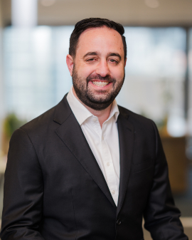 A man smiling, wearing a black suit and white shirt, posing for a headshot.
