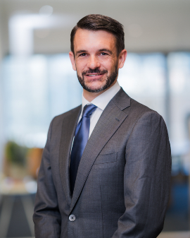 A man smiling, wearing a grey suit and blue tie, posing for a headshot.