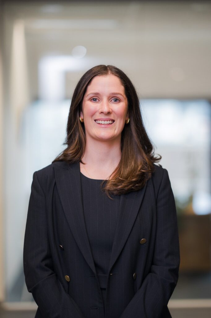 A women smiling, dressed in a black blazer, posing for a headshot.