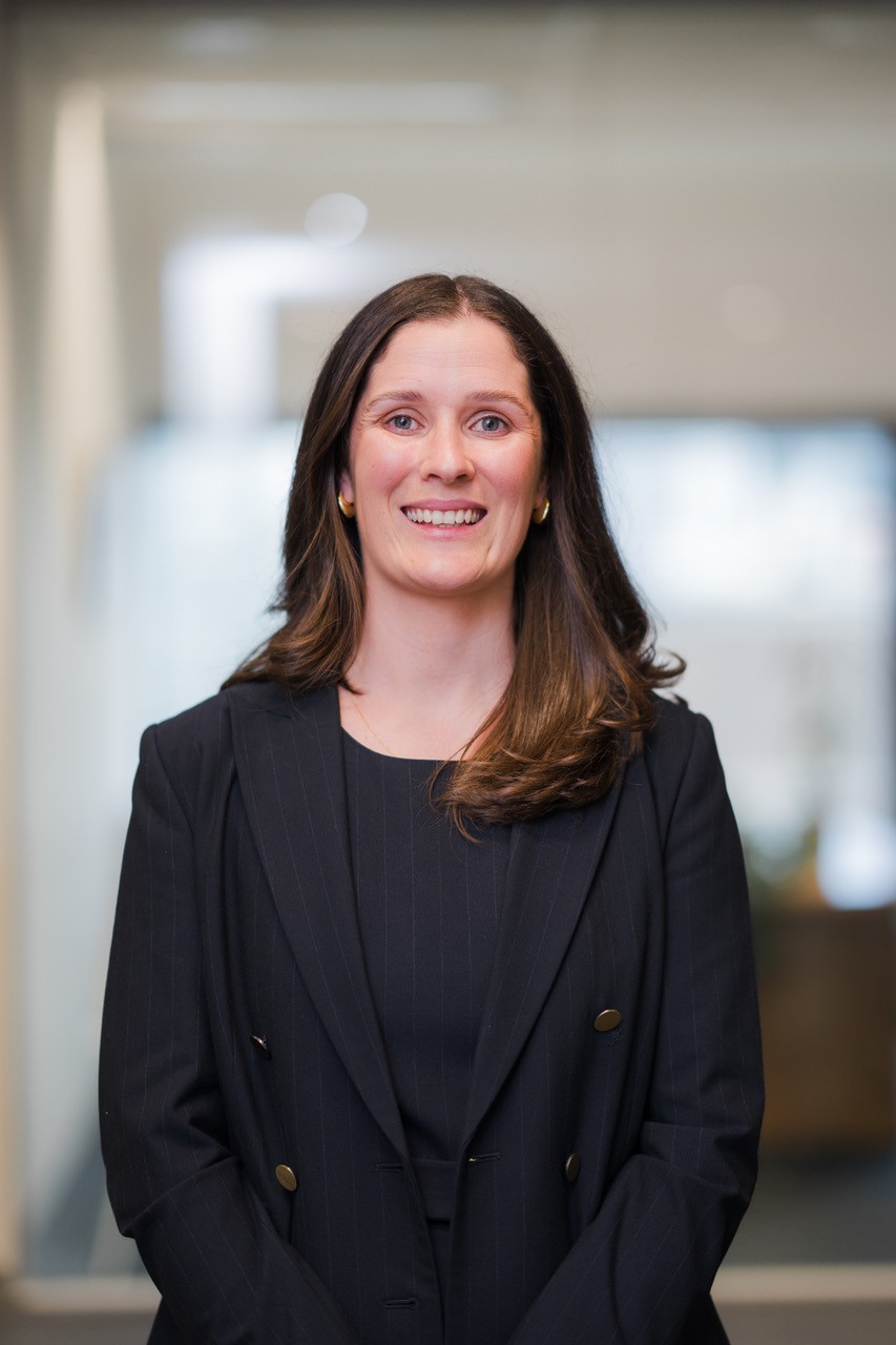 A women smiling, dressed in a black blazer, posing for a headshot.